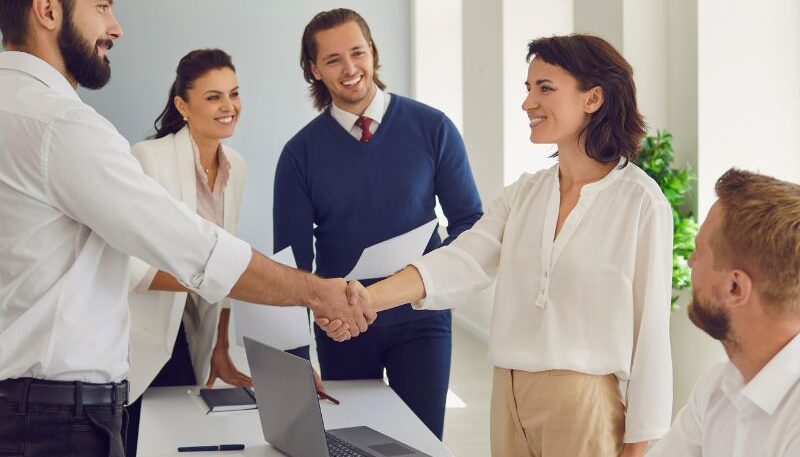 Five people in a boardroom shaking hands after negotiating a supplier arrangement through managed procurement services.